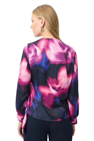 A woman shown from the back wearing a long-sleeve blouse with a vibrant abstract floral print in pink, purple, and blue over a dark navy base, paired with dark navy pants against a light gray studio background.