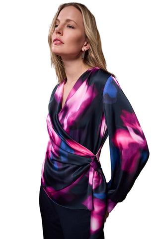 A woman wearing a long-sleeve wrap-style top with a bold abstract print in pink, purple, and blue over a dark base, posed against a warm brown curtain backdrop.