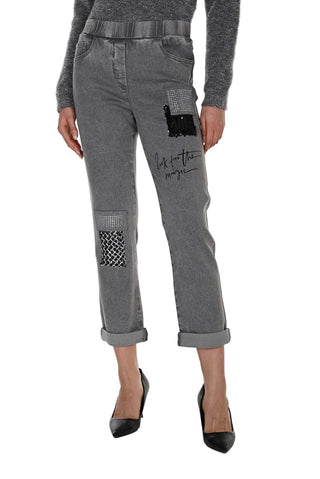 A model from the waist down wearing gray patchwork cropped jeans with black and white patterned pocket patches and script embroidery on the thigh, paired with black heels.