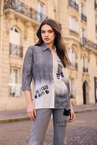 A woman stands on a cobblestone street in front of a beige stone building, wearing a mixed-media shirt with vertical gray and white stripes fading into a white graphic print, paired with gray pants.
