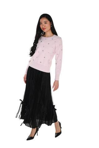 Woman wearing a light pink sweater with decorative elements and a black skirt on a white background