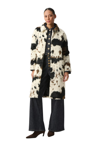 A model stands against a light gray studio background wearing a long black-and-cream cow-print coat over a dark button-front top, wide-leg dark jeans, and dark pointed shoes.