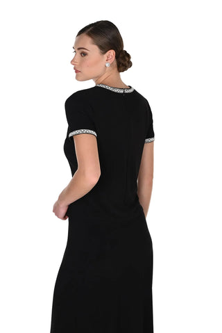 A woman modeling a black short-sleeve maxi dress from the back, featuring a decorative silver and black patterned trim along the neckline and sleeve cuffs, set against a plain white studio background.