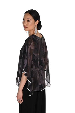 A woman wearing a sheer black floral-patterned cape-style top with white piping trim, shown from a side-back angle against a plain white studio background.