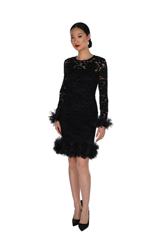 A woman modeling a black lace long-sleeve mini dress with feathered trim at the hem and cuffs, paired with black pointed-toe heels, against a plain white studio background.