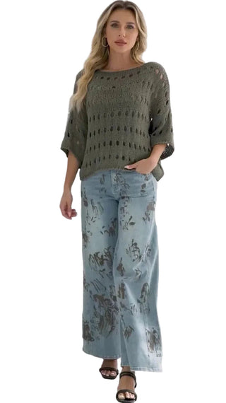 Woman wearing a green sweater and light blue floral pants in a room with a black chair and white walls.