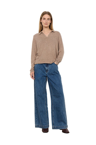 A woman stands against a plain white wall wearing a beige long-sleeve knit sweater with a V-neck and wide-leg blue jeans.