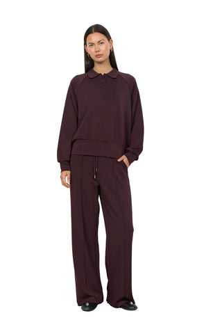 A model stands against a plain white wall wearing a matching dark plum-burgundy loungewear set consisting of a long-sleeve collared top and wide-leg drawstring pants.