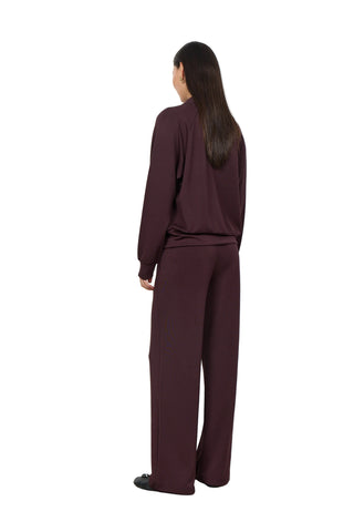 A woman stands with her back to the camera wearing a matching dark plum-burgundy lounge set consisting of a long-sleeve top and wide-leg trousers, set against a plain white wall and light concrete floor.