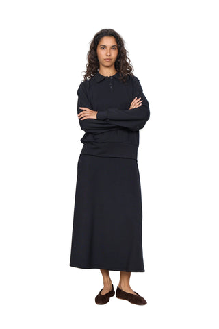 A woman stands with arms crossed wearing a black long-sleeve polo-style top and a matching black midi skirt, paired with dark brown flat shoes, against a plain white studio background.