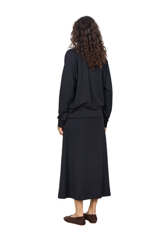 A woman with long curly brown hair is shown from behind wearing a black long-sleeve top and a matching black midi skirt, paired with dark brown flat shoes, against a plain white wall.