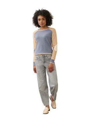 A model stands in a minimalist studio setting wearing a color-blocked raglan long-sleeve top with a dusty blue body and beige sleeves, paired with light-wash jeans and beige loafers.