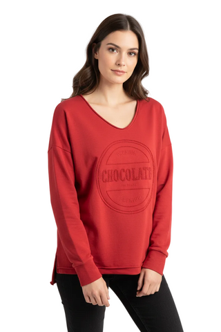 A woman wearing a red long-sleeve V-neck sweatshirt with a tonal circular 'CHOCOLATE' graphic on the front, paired with black pants, against a light gray studio background.