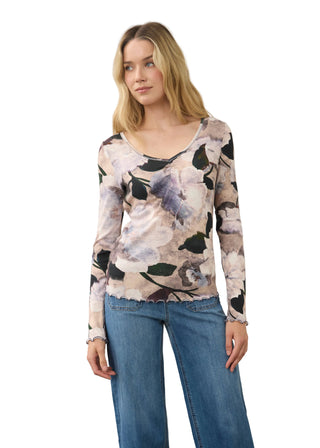 A woman modeling a long-sleeve scoop-neck top with a muted floral print in beige, gray, black, and lavender, paired with blue jeans, against a plain light gray studio background.