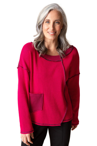 A smiling woman with gray hair wearing a bright magenta waffle-knit asymmetrical sweater with a front pocket, paired with black pants, against a solid beige background.