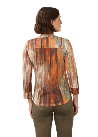 A woman viewed from the back wearing a long-sleeve button-up shirt with a vertical multicolor abstract print in orange, brown, beige, and teal, paired with olive green pants, against a plain light gray background.