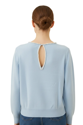 A woman viewed from the back wearing a light blue knit sweater with a pearl-trimmed keyhole cutout at the neckline, paired with blue jeans against a plain cream background.