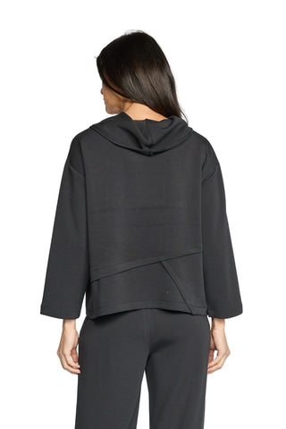 A woman viewed from the back wearing a dark charcoal gray hooded long-sleeve top and matching dark pants against a light gray studio background.