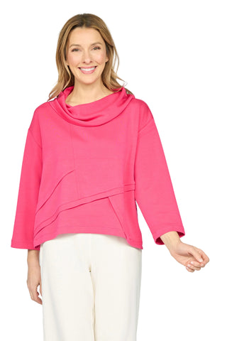 A woman modeling a bright pink cowl-neck top with layered asymmetrical front panels and three-quarter sleeves, paired with white pants, against a light gray studio background.