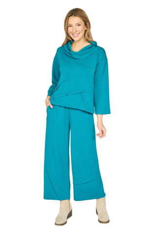 A woman modeling a matching teal two-piece outfit consisting of a cowl-neck long-sleeve top and wide-leg cropped pants, paired with beige ankle boots, against a light gray studio background.