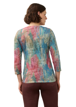 A woman viewed from the back wearing a three-quarter sleeve top with an abstract painterly pattern in blue, pink, and green, paired with dark brown pants, against a plain light background.