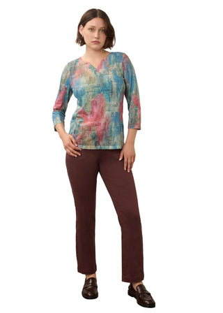 A woman modeling a three-quarter sleeve V-neck top with an abstract multicolor print in shades of teal, pink, and blue, paired with dark brown trousers and brown leather loafers, standing against a plain off-white wall.