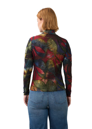 A woman viewed from the back wearing a long-sleeve mock-neck top with a complex multicolor abstract pattern in red, green, blue, and yellow, paired with blue jeans, against a plain light gray background.