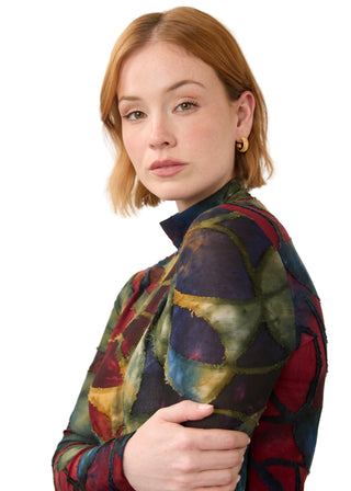A woman with short auburn hair wearing a colorful patchwork-patterned long-sleeve top and small gold hoop earrings, photographed against a plain light gray studio background.