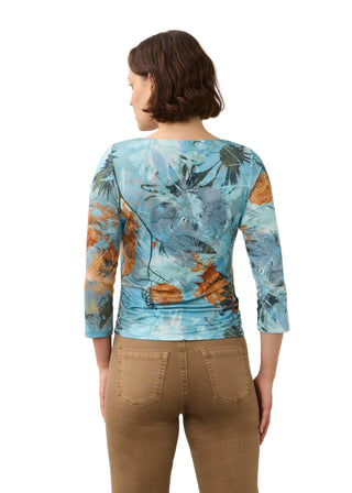 A woman viewed from the back wearing a light blue three-quarter sleeve top with an abstract botanical print in shades of blue, brown, and gray, paired with brown pants, against a plain light beige background.