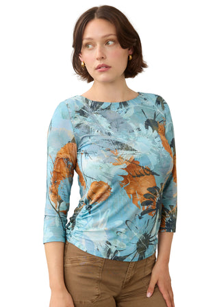 A woman wearing a light blue three-quarter sleeve top with an abstract botanical print in orange, black, and white, paired with brown pants, against a plain light gray background.
