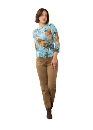 A woman modeling a light blue tropical print long-sleeve top paired with brown pants and dark brown ankle boots, standing against a plain off-white wall and concrete floor.