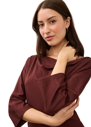 A woman with dark brown hair wearing a draped, three-quarter sleeve burgundy top, posing against a plain light gray background.