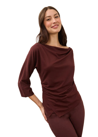 A woman modeling a draped, three-quarter sleeve top and matching leggings in a deep burgundy color, set against a plain light gray studio background.