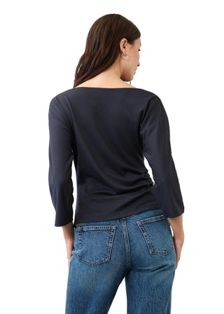 A woman viewed from the back wearing a dark navy long-sleeve boat-neck top and blue jeans, standing against a plain light gray studio background.