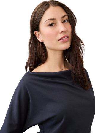A woman with long brown hair wearing a dark navy blue off-the-shoulder top and small hoop earrings, photographed against a clean light gray background.