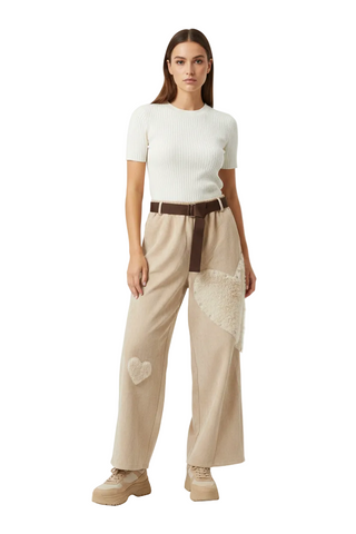 A model stands against a light gray studio background wearing a white short-sleeve ribbed top, beige wide-leg pants with a brown belt and cream lace/patch details, and beige platform sneakers.