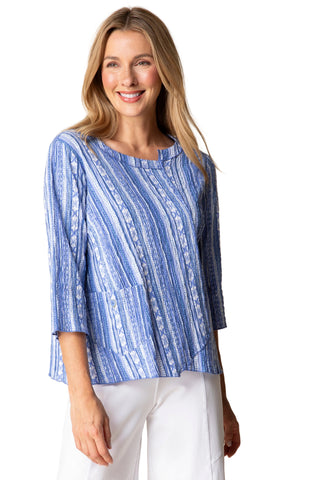 Woman wearing a blue patterned top and white pants against a beige background