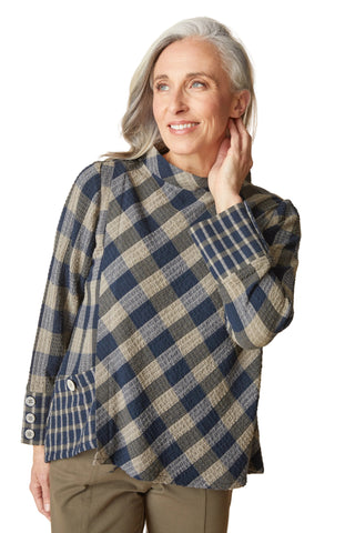 An older woman with gray hair is wearing a blue, gray, and beige plaid long-sleeve top with a mock neck and buttoned cuffs, paired with olive green pants, against a solid beige background.