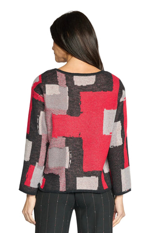 A woman viewed from the back wearing a cropped knit sweater with a bold geometric block pattern in red, black, and gray, paired with black pinstriped trousers against a light gray studio background.