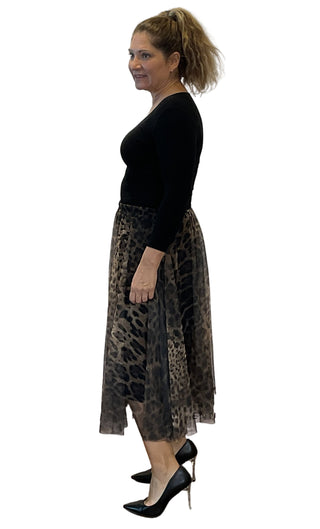Woman wearing a black top and leopard print skirt standing against a white wall with visible branding.