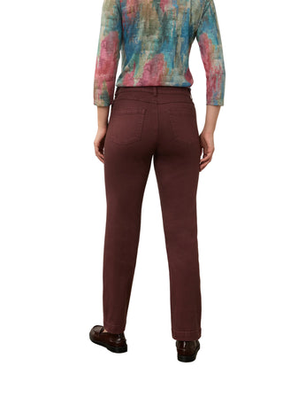 A person shown from the back wearing slim-fit burgundy trousers, a multicolored patterned long-sleeve top, and dark brown leather shoes, standing against a plain off-white wall and concrete floor.