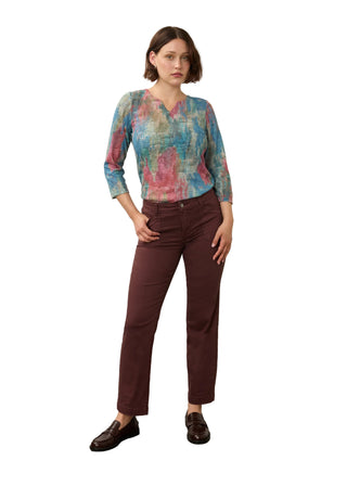 A model stands in a studio wearing a long-sleeve V-neck blouse with a blue, pink, and green abstract print, paired with dark brown trousers and dark brown leather loafers.
