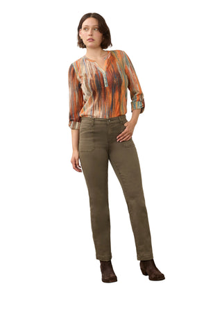 A woman modeling a long-sleeve button-up shirt with an abstract orange, brown, and beige pattern, paired with olive green trousers and dark brown ankle boots, standing against a neutral studio backdrop.