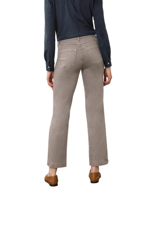 A person shown from the back wearing light gray cropped jeans, a dark navy long-sleeve shirt, and brown shoes in a studio setting with a plain off-white wall and textured concrete floor.