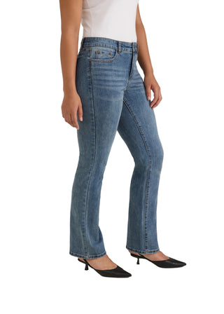 A model wearing a pair of medium-wash blue denim flare jeans, styled with a white sleeveless top and black pointed-toe mule heels, photographed against a neutral light gray studio background.
