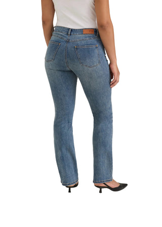 Rear view of a model wearing medium-wash blue denim jeans with a straight-to-flare leg, paired with a white top and black pointed-toe mule heels, against a light beige studio background.