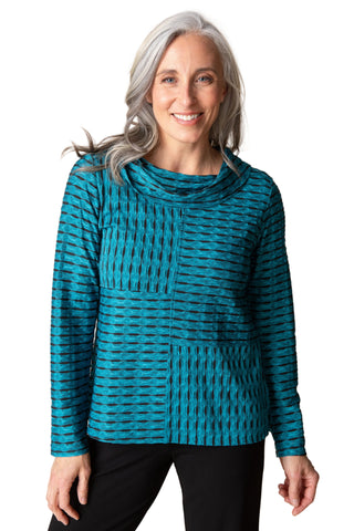 A smiling woman with gray hair wearing a teal and black patterned long-sleeve top with a cowl neck, paired with black pants, against a solid beige background.