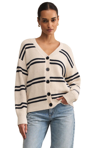 Woman wearing a cream cardigan with black stripes and buttons, paired with light blue jeans on a beige background.