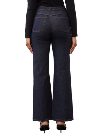 Rear view of a model wearing dark blue wide-leg jeans with a black long-sleeve top and black heels, standing on a concrete floor against a white studio background.