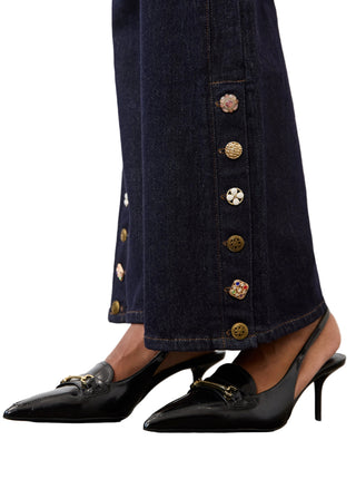 Close-up of a person wearing dark navy wide-leg jeans with decorative gold-tone buttons down the side and black pointed slingback heels with gold hardware, standing on a wooden floor against a light wall.